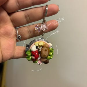 K&ouml;nnte beinhalten: Ein kleiner, runder, silberfarbener Spiegelanh&auml;nger mit einem Klappdeckel. Der Anh&auml;nger zeigt eine farbenfrohe, skulpturale Szene mit einem Marienk&auml;fer, einem B&auml;ren und Blumen. Der Anh&auml;nger ist an einem silbernen Schl&uuml;sselring mit rosa Perlen befestigt.