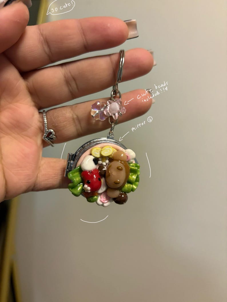 K&ouml;nnte beinhalten: Ein kleiner, runder, silberfarbener Spiegelanh&auml;nger mit einem Klappdeckel. Der Anh&auml;nger zeigt eine farbenfrohe, skulpturale Szene mit einem Marienk&auml;fer, einem B&auml;ren und Blumen. Der Anh&auml;nger ist an einem silbernen Schl&uuml;sselring mit rosa Perlen befestigt.