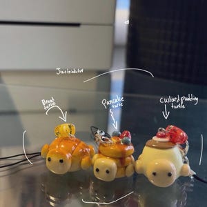 Op de afbeelding: Drie miniatuur schildpadhangers, elk gemaakt om op een ander dessert te lijken: brood, pannenkoeken en custardpudding. De hangers zijn geel, oranje en bruin, met kleine details zoals bessen en siroop. Het woord "Yummy!" staat erboven.