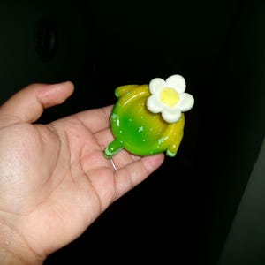 Può includere: Un oggetto decorativo a forma di tartaruga, verde e giallo, ornato da un fiore bianco e giallo. L'oggetto lucido è tenuto in mano e potrebbe essere una spilla o un fermaglio.