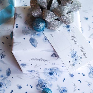 May include: A white gift box wrapped in blue floral patterned paper with a silver bow and blue ornaments. The gift tag says "Happy Holidays" and has a blank space for "from:" and "to:".