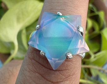 Puede incluir: Un anillo grande, iridiscente en forma de estrella, con una banda plateada. La estrella tiene un degradado de color azul claro y verde. El anillo se encuentra sobre un fondo de follaje verde.
