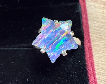 Puede incluir: Un anillo en forma de estrella con una gema iridiscente grande. La gema muestra una gama de colores, incluyendo azul, verde y púrpura. La banda del anillo es plateada, y la gema está sujeta por cuatro garras plateadas.