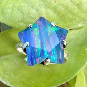 Puede incluir: Un anillo en forma de estrella con una gran gema azul facetada. La gema tiene cualidades iridiscentes con toques de verde y morado. La banda y las garras del anillo son de color plateado, sobre una hoja verde.