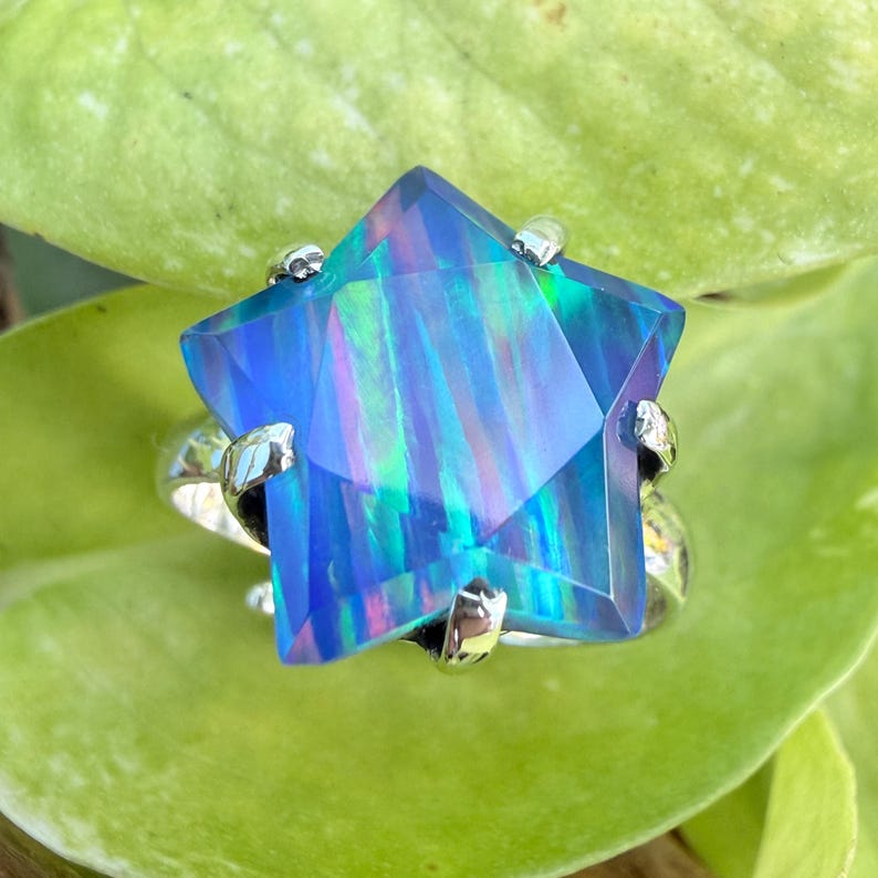 Puede incluir: Un anillo en forma de estrella con una gran gema azul facetada. La gema tiene cualidades iridiscentes con toques de verde y morado. La banda y las garras del anillo son de color plateado, sobre una hoja verde.