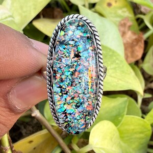 Op de afbeelding: Een ovale ring met een zilverkleurige band en een kleurrijke, mozaïekachtige steen. De steen heeft een mix van blauwe, groene, oranje en zwarte fragmenten, wat een levendig ontwerp creëert. De ring staat tegen een achtergrond van groen gebladerte.
