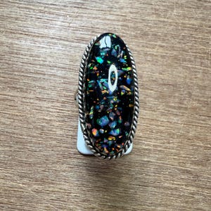 Op de afbeelding: Ovale ring met een zwarte steen ingelegd met kleurrijke iriserende fragmenten. De ring heeft een zilverkleurige, gedraaide, touwachtige rand. De ring is geplaatst op een wit label, rustend op een houten oppervlak.
