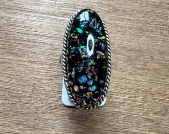 May include: Oval-shaped ring with a black stone inlaid with colorful iridescent fragments. The ring has a silver-colored, twisted rope-like border. The ring is set on a white tag, resting on a wooden surface.