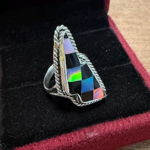 May include: A silver ring with a triangular, mosaic-style gemstone. The gemstone features a checkerboard pattern of black, iridescent, and colorful squares. The ring is set in a dark red velvet box.