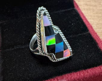 Puede incluir: Un anillo de plata con una piedra preciosa triangular de estilo mosaico. La piedra preciosa presenta un patrón de tablero de ajedrez de cuadrados negros, iridiscentes y coloridos. El anillo está en una caja de terciopelo rojo oscuro.
