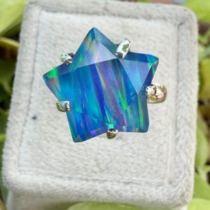 Puede incluir: Un anillo en forma de estrella con una gran piedra preciosa de &oacute;palo azul facetado. El &oacute;palo muestra tonos de azul y verde. La banda y las garras del anillo son de color plateado. El anillo est&aacute; sobre una superficie gris texturizada.