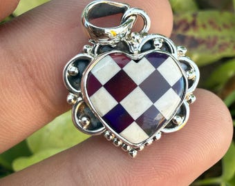 Könnte beinhalten: Silberanhänger mit herzförmigem Design. Das Herz hat ein Schachbrettmuster aus weißen, weinroten und violetten Quadraten. Der Anhänger hat einen dekorativen Silberrand und eine Schlaufe für eine Kette.