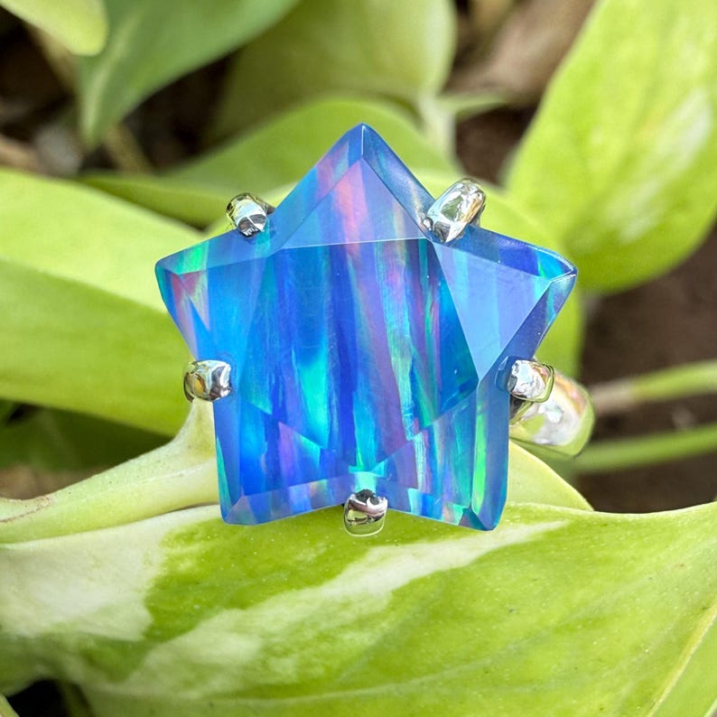 Puede incluir: Un anillo en forma de estrella con una gran piedra preciosa azul facetada. La piedra preciosa tiene una calidad vibrante e iridiscente con vetas azules, verdes y moradas. La banda del anillo es plateada y la piedra preciosa est&aacute; sujeta por garras plateadas.