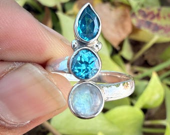 Könnte beinhalten: Ein Silberring mit drei Edelsteinen: einem tropfenförmigen blauen Edelstein, einem runden blauen Edelstein und einem runden, irisierenden Mondstein. Der Ring wird vor einem Hintergrund aus grünen Blättern gehalten.