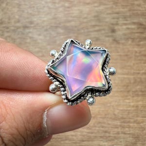 Puede incluir: Un anillo de plata con una piedra preciosa en forma de estrella con colores iridiscentes. La piedra preciosa tiene un diseño facetado, y el anillo tiene un engaste detallado y texturizado con pequeños acentos redondos. El anillo está sostenido por los dedos de una persona.
