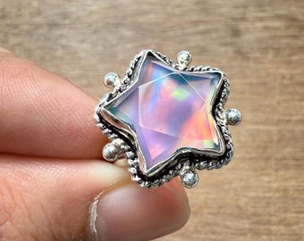 Puede incluir: Un anillo de plata con una piedra preciosa en forma de estrella con colores iridiscentes. La piedra preciosa tiene un diseño facetado, y el anillo tiene un engaste detallado y texturizado con pequeños acentos redondos. El anillo está sostenido por los dedos de una persona.