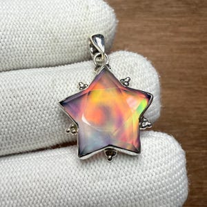 Puede incluir: Un colgante en forma de estrella con un marco plateado. El centro del colgante presenta una piedra iridiscente colorida con tonos naranja, amarillo y verde. El colgante está suspendido de un lazo plateado.