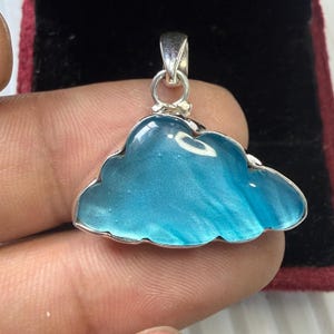 May include: A cloud-shaped pendant with a silver-colored frame. The pendant has a light blue, translucent center with white streaks. The bail is also silver-colored. The pendant is likely made of glass or resin.