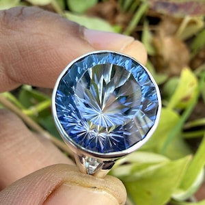 Op de afbeelding: Zilveren ring met een grote, gefacetteerde blauwe edelsteen. De edelsteen is helder, lichtblauw, geslepen om een sterrenuitbarstingseffect te creëren. De ring staat tegen een vage groene achtergrond.