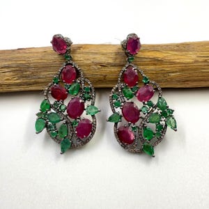 Könnte beinhalten: Ein Paar verzierte Ohrhänger mit oval geschliffenen Rubin- und Smaragd-Edelsteinen. Die Ohrringe haben ein dunkles Metall-Filigran-Design mit kleinen Akzentsteinen und werden auf einer Holzoberfläche präsentiert.