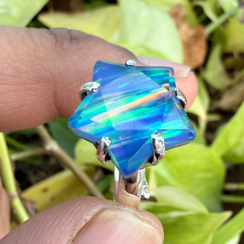 Puede incluir: Anillo de plata con una gran gema en forma de estrella, que muestra colores azules, verdes y amarillos. La gema est&aacute; sujeta por seis garras de plata. El anillo se encuentra sobre un fondo de follaje verde. La gema est&aacute; facetada.