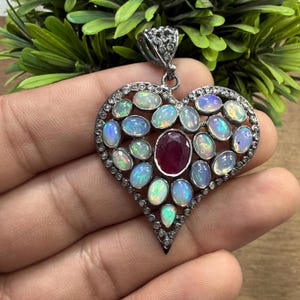 May include: Heart-shaped pendant with a central oval ruby surrounded by opals and small clear stones. The pendant is made of silver-colored metal and has a bail at the top for a chain.