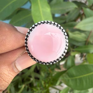 May include: A round ring featuring a smooth, polished, light pink cabochon stone set in a decorative, oxidized silver-toned metal frame with a beaded edge. The setting is circular and highlights the soft, opaque pink color of the gemstone.