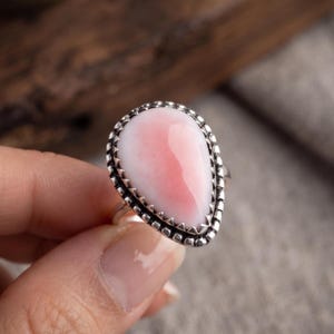 May include: A teardrop-shaped ring featuring a smooth, polished pink and white stone set in a textured silver metal bezel. The setting has a decorative, notched edge that frames the stone, which displays a gradient of soft pink to white.