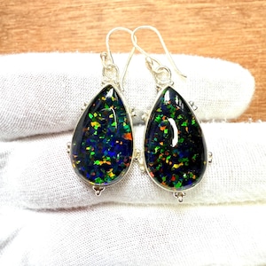 May include: A pair of silver drop earrings featuring teardrop-shaped black opal stones. The opals display a vibrant play of color with green, blue, and orange flashes. The earrings have a simple hook design and are set in a silver bezel.