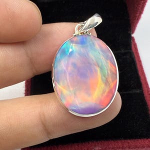 Könnte beinhalten: Ovale Opal-Anhänger mit einer silbernen Öse. Der Opal zeigt ein lebendiges Farbspiel, darunter Farbtöne von Blau, Grün, Gelb, Orange und Lila. Der Anhänger wird von einer Hand gehalten, die die schillernde Qualität des Edelsteins zeigt.