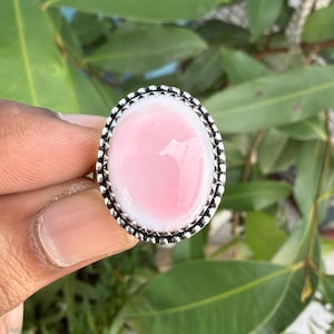 May include: An oval-shaped ring featuring a smooth, light pink cabochon stone set in a decorative silver-toned metal frame with a scalloped edge. The setting has a textured, beaded border surrounding the central pink stone.