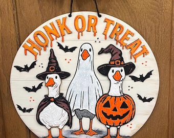 May include: Round wooden Halloween sign with the words "HONK OR TREAT" in orange dripping letters. The sign features three geese dressed in Halloween costumes: a witch, a ghost, and a jack-o'-lantern. Black bats are scattered around the geese.