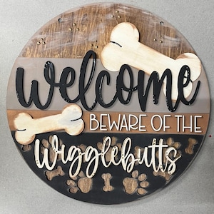 以下が含まれることがあります： 「Welcome Beware of the Wigglebutts」の文字が入った丸い木製サイン。犬の骨と足跡のデザインが施されています。茶色、黒、グレーの色合いで、多層構造になっています。