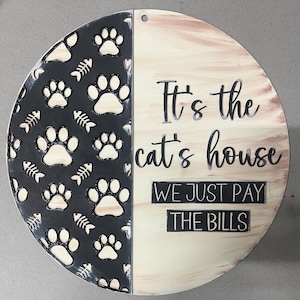May include: A circular wooden sign, divided into two halves. One side is black with paw prints and fish bones, the other is cream-coloured with the text "It's the cat's house, we just pay the bills."