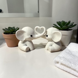May include: Two white elephant figurines with black eyes and large ears. Their trunks form a heart shape. The elephants sit on a white surface, with potted succulents on either side. A white towel is folded in the foreground.