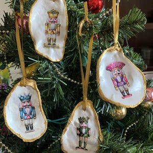 May include: Christmas tree ornaments featuring nutcracker designs. Each ornament is made from a white oyster shell with a gold-painted edge and a gold ribbon for hanging. The nutcrackers are depicted in various colors.