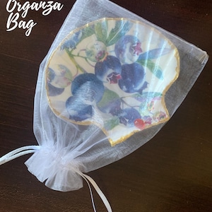 K&ouml;nnte beinhalten: Eine dekorative, muschelf&ouml;rmige Schale mit Blaubeer-Design, pr&auml;sentiert in einem wei&szlig;en Organzabeutel. Die Schale hat einen goldenen Rand und eine detaillierte Illustration von Blaubeeren und Bl&auml;ttern. Der Text "Includes Organza Bag" ist sichtbar.