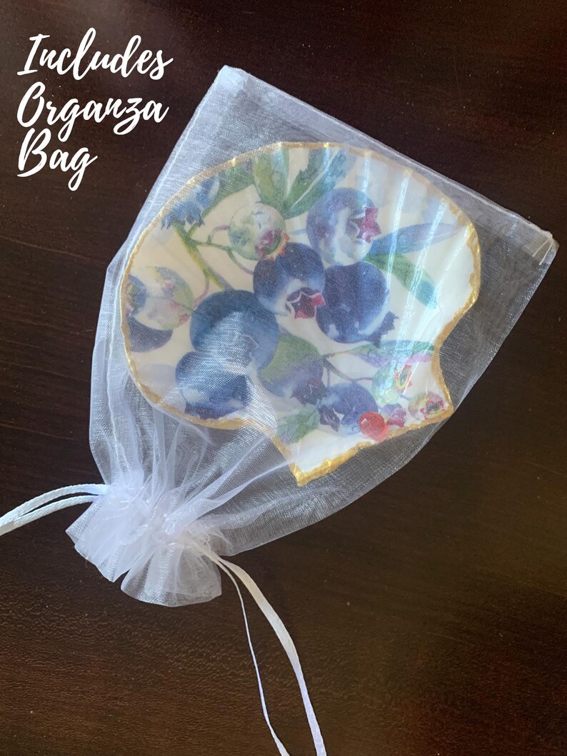 K&ouml;nnte beinhalten: Eine dekorative, muschelf&ouml;rmige Schale mit Blaubeer-Design, pr&auml;sentiert in einem wei&szlig;en Organzabeutel. Die Schale hat einen goldenen Rand und eine detaillierte Illustration von Blaubeeren und Bl&auml;ttern. Der Text "Includes Organza Bag" ist sichtbar.