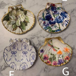 K&ouml;nnte beinhalten: Vier dekorative Muschelschalen f&uuml;r Schmuck. Jede Schale ist mit einem einzigartigen Design bemalt und mit Gold verziert. Zu den Designs geh&ouml;ren ein Blumenmuster, Blaubeeren, ein blau-wei&szlig;es Muster und eine Libelle mit Blumen. Jede Schale hat einen Perlenakzent.
