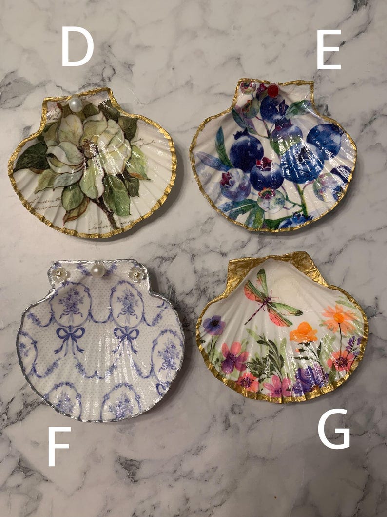 K&ouml;nnte beinhalten: Vier dekorative Muschelschalen f&uuml;r Schmuck. Jede Schale ist mit einem einzigartigen Design bemalt und mit Gold verziert. Zu den Designs geh&ouml;ren ein Blumenmuster, Blaubeeren, ein blau-wei&szlig;es Muster und eine Libelle mit Blumen. Jede Schale hat einen Perlenakzent.