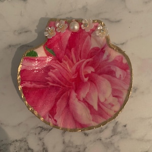K&ouml;nnte beinhalten: Dekorative Muschelschalen-Schmuckschale. Die Schale zeigt ein lebendiges rosa Blumendesign mit einem goldfarbenen Rand. Kleine wei&szlig;e Bl&uuml;ten und eine Perle akzentuieren den oberen Rand und verleihen einen Hauch von Eleganz.