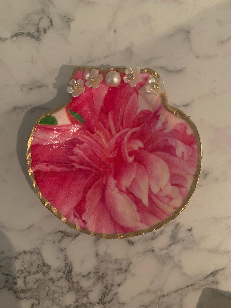 K&ouml;nnte beinhalten: Dekorative Muschelschalen-Schmuckschale. Die Schale zeigt ein lebendiges rosa Blumendesign mit einem goldfarbenen Rand. Kleine wei&szlig;e Bl&uuml;ten und eine Perle akzentuieren den oberen Rand und verleihen einen Hauch von Eleganz.