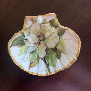 May include: A decorative seashell with a painted magnolia flower design. The shell is white with a gold-painted edge and a pearl accent. The magnolia has white and yellow petals with green leaves.