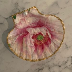 K&ouml;nnte beinhalten: Dekorative Muschelschalen-Schmuckschale. Die Schale ist mit einem rosa-wei&szlig;en Blumenmuster und einem goldfarbenen Rand bemalt. Eine kleine Perle ist am oberen Rand befestigt. Die Schale ist f&uuml;r kleine Gegenst&auml;nde gedacht.