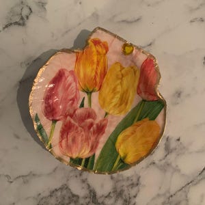 K&ouml;nnte beinhalten: Eine dekorative, unregelm&auml;&szlig;ig geformte Platte mit rosa Hintergrund und einem floralen Tulpenmuster. Die Tulpen sind in Gelb-, Orange- und Rosat&ouml;nen gehalten, mit gr&uuml;nen Bl&auml;ttern. Der Rand ist goldfarben.
