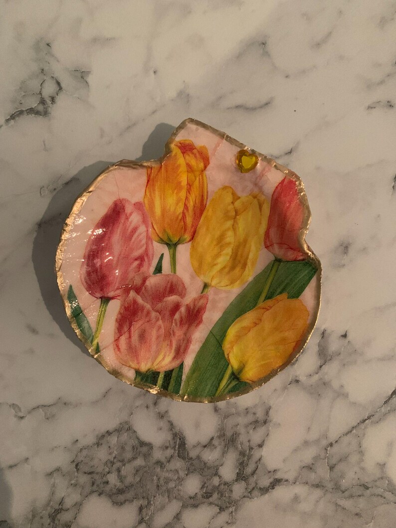 K&ouml;nnte beinhalten: Eine dekorative, unregelm&auml;&szlig;ig geformte Platte mit rosa Hintergrund und einem floralen Tulpenmuster. Die Tulpen sind in Gelb-, Orange- und Rosat&ouml;nen gehalten, mit gr&uuml;nen Bl&auml;ttern. Der Rand ist goldfarben.