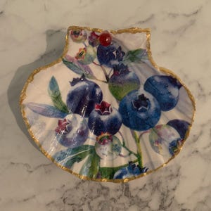 K&ouml;nnte beinhalten: Dekorative muschelf&ouml;rmige Schale mit wei&szlig;em Innenraum und Blaubeer-Design. Die Blaubeeren sind in verschiedenen Blaut&ouml;nen mit gr&uuml;nen Bl&auml;ttern gehalten. Der Rand ist mit Goldfarbe verziert, und eine rote Perle ist angebracht.