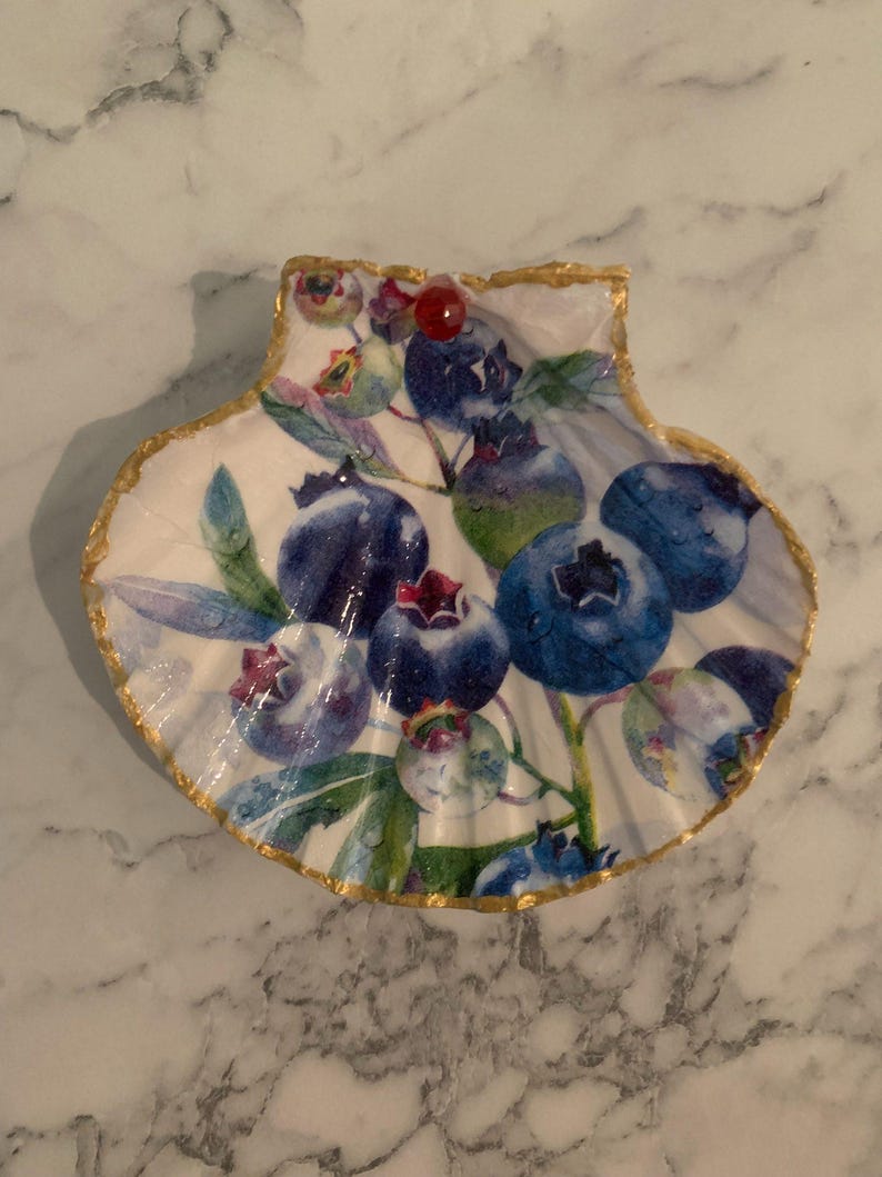 K&ouml;nnte beinhalten: Dekorative muschelf&ouml;rmige Schale mit wei&szlig;em Innenraum und Blaubeer-Design. Die Blaubeeren sind in verschiedenen Blaut&ouml;nen mit gr&uuml;nen Bl&auml;ttern gehalten. Der Rand ist mit Goldfarbe verziert, und eine rote Perle ist angebracht.