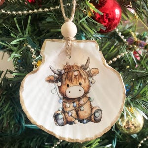 May include: A seashell Christmas ornament featuring a Highland cow illustration. The cow is brown and tan, adorned with a string of lights with star-shaped accents. The shell has a gold-painted edge and is suspended by a jute cord and wooden bead.