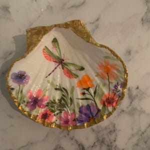 K&ouml;nnte beinhalten: Handbemalte Muschelschale mit goldfarbenem Rand. Das Innere zeigt eine Libelle und bunte Blumen in Rosa-, Lila- und Oranget&ouml;nen. Die Schale ist wei&szlig; und liegt auf einer Marmoroberfl&auml;che.
